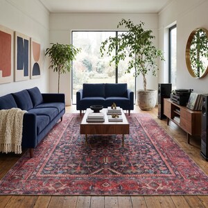 May include: A living room featuring a large rectangular area rug with a dark red and navy blue intricate pattern, two navy blue sofas, a wooden coffee table with books, a wooden media console, and a round gold-framed mirror on the wall.