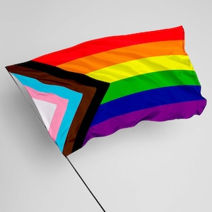 Könnte beinhalten: Eine Pride-Flagge mit einem schwarzen und braunen Dreieck auf der linken Seite, das Menschen mit Farbe repräsentiert, und einem rosa, blauen und weißen Dreieck auf der rechten Seite, das Transgender-Menschen repräsentiert. Die Flagge ist aus Stoff gefertigt und weht im Wind.