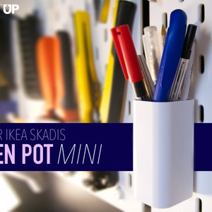 May include: White plastic pen holder designed for IKEA SKADIS pegboard. The holder is labelled "PEN POT MINI". The holder is filled with pens in red, blue and black.