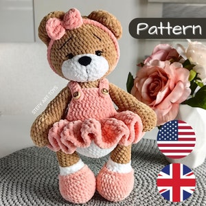 Può includere: Orso di peluche lavorato a uncinetto con un vestito rosa, una fascia marrone e un fiocco rosa. L'orso ha occhi bianchi e un naso bianco. L'orso è seduto su una superficie bianca e grigia. Il testo "Pattern" è nell'angolo in alto a destra dell'immagine. L'immagine include anche le bandiere americana e britannica.