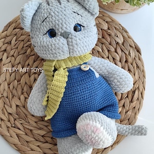 Könnte beinhalten: Ein graues gehäkeltes Katzenspielzeug, das einen gelben Schal und eine blaue Latzhose trägt. Die Katze hat blaue Augen und rosa Pfoten. Das Spielzeug sitzt auf einer geflochtenen Weidenoberfläche.