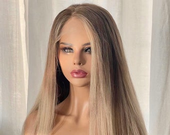 Puede incluir: Una peluca larga y en capas con una parte delantera de encaje, que presenta una mezcla de reflejos rubios y marrones claros. La peluca está peinada en una cabeza de maniquí, mostrando su línea de cabello de aspecto natural y mechones lisos y fluidos.