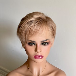 Puede incluir: Una peluca corta rubia con raya lateral y estilo en capas. La peluca está peinada con raya lateral y tiene un aspecto natural y despeinado.