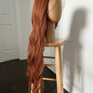 May include: A long, wavy, auburn wig is displayed on a mannequin head. The wig is styled with a lace front and is positioned on a wooden stool. The hair cascades down, showcasing its length and texture. The background is a white wall and dark wood floor.