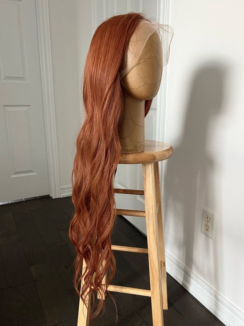 May include: A long, wavy, auburn wig is displayed on a mannequin head. The wig is styled with a lace front and is positioned on a wooden stool. The hair cascades down, showcasing its length and texture. The background is a white wall and dark wood floor.