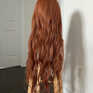 May include: A long, wavy auburn wig is displayed on a wooden stool against a white wall. The wig has a natural-looking hairline and a voluminous, curly texture. The hair cascades down in loose waves, creating a full and flowing appearance.