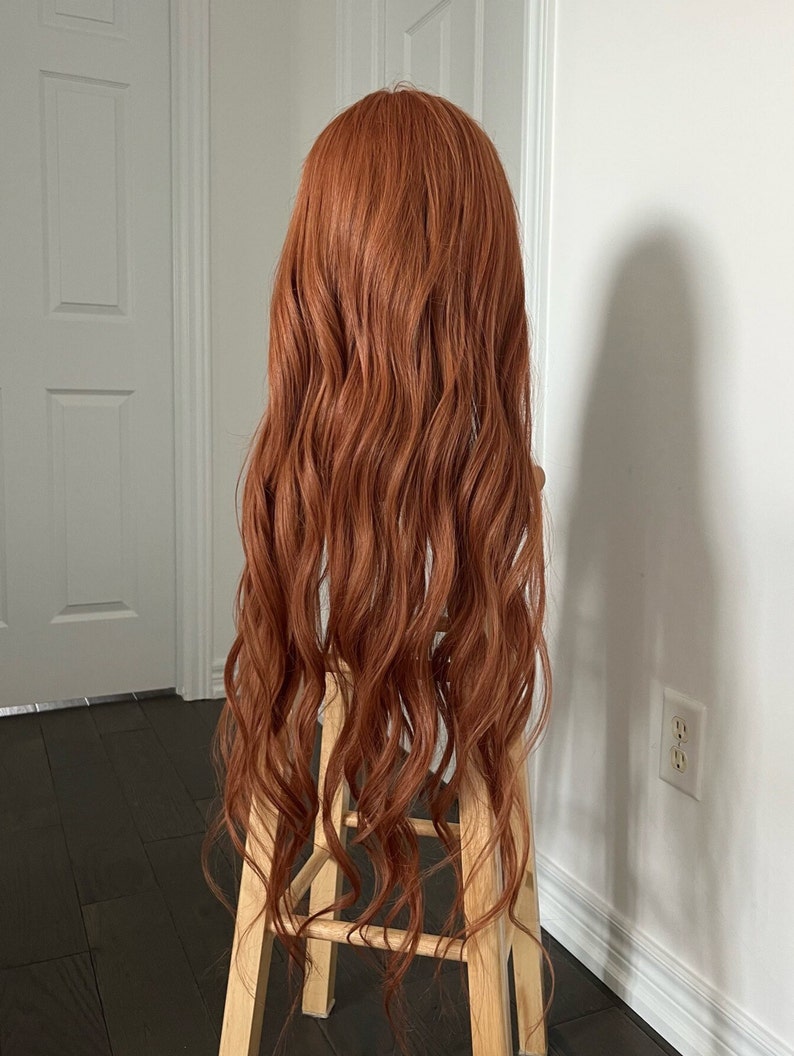 May include: A long, wavy auburn wig is displayed on a wooden stool against a white wall. The wig has a natural-looking hairline and a voluminous, curly texture. The hair cascades down in loose waves, creating a full and flowing appearance.