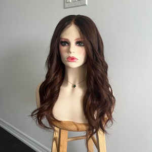 Op de afbeelding: Een bruin golvend pruik op een mannequinhoofd met een groene klavertjesvier-ketting. De mannequin zit op een houten kruk.