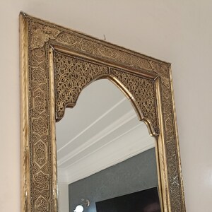 May include: Ornate gold-colored mirror with an arched top. The frame features intricate, detailed patterns. The mirror reflects a room with a television and a wall-mounted light fixture. Decorative wall mirror.