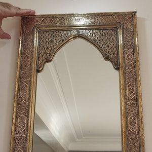 May include: Ornate rectangular mirror with a gold-colored frame featuring intricate, dark-colored patterns. The top of the mirror has an arched design. The mirror is being held up against a white wall.