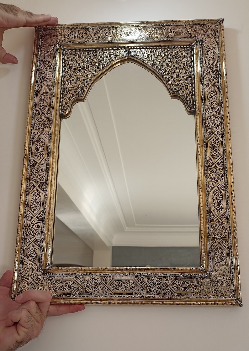 May include: Ornate rectangular mirror with a gold-colored frame featuring intricate, dark-colored patterns. The top of the mirror has an arched design. The mirror is being held up against a white wall.