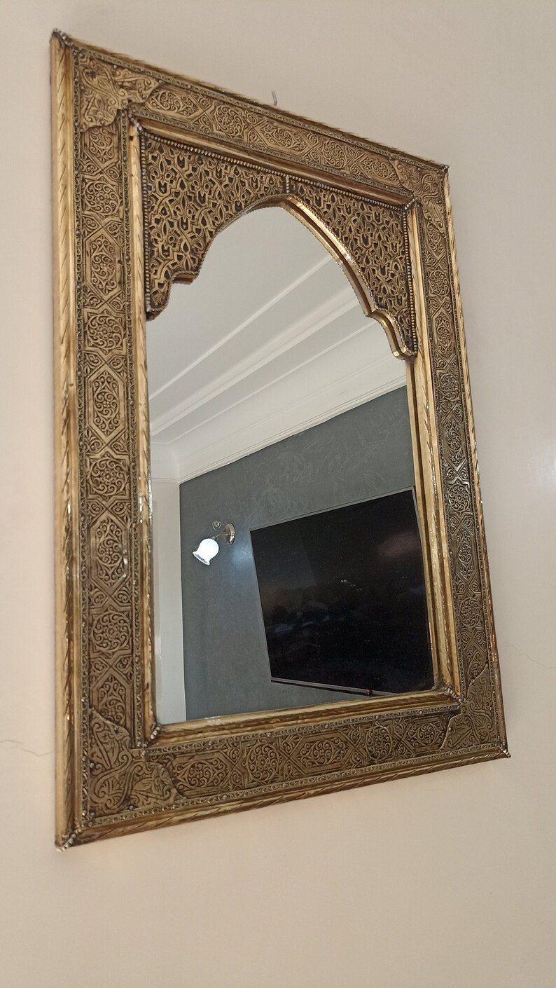 May include: Ornate gold-colored mirror with an arched top. The frame features intricate, detailed patterns. The mirror reflects a room with a television and a wall-mounted light fixture. Decorative wall mirror.
