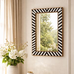 Peut inclure: Un miroir mural rectangulaire avec un cadre à chevrons noir et blanc et une bordure dorée. Le miroir est accroché à un mur blanc. Un vase de fleurs blanches est placé devant une fenêtre avec des rideaux crème.