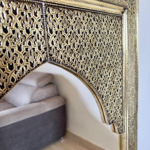 May include: Ornate gold-colored mirror with intricate, geometric patterns. The mirror's frame features detailed relief work and a dark background, creating a striking contrast. The design is reminiscent of traditional Moroccan craftsmanship.