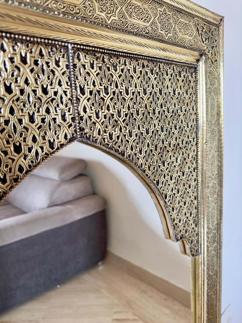 May include: Ornate gold-colored mirror with intricate, geometric patterns. The mirror's frame features detailed relief work and a dark background, creating a striking contrast. The design is reminiscent of traditional Moroccan craftsmanship.