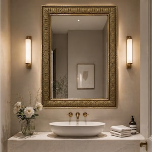 Peut inclure: Intérieur de salle de bain avec un grand miroir rectangulaire à cadre doré orné. Au-dessus d'un lavabo ovale blanc avec robinetterie dorée, deux appliques murales verticales encadrent le miroir. Un vase de fleurs blanches est posé sur le comptoir.