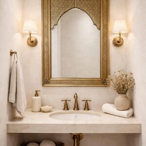 Intérieur de salle de bain avec miroir doré, appliques murales et lavabo. La vanité comprend un panier en osier avec des serviettes roulées, un distributeur de savon et un vase avec des fleurs séchées. La palette de couleurs est neutre avec des accents dorés.