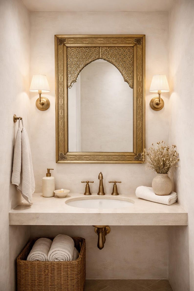 Intérieur de salle de bain avec miroir doré, appliques murales et lavabo. La vanité comprend un panier en osier avec des serviettes roulées, un distributeur de savon et un vase avec des fleurs séchées. La palette de couleurs est neutre avec des accents dorés.