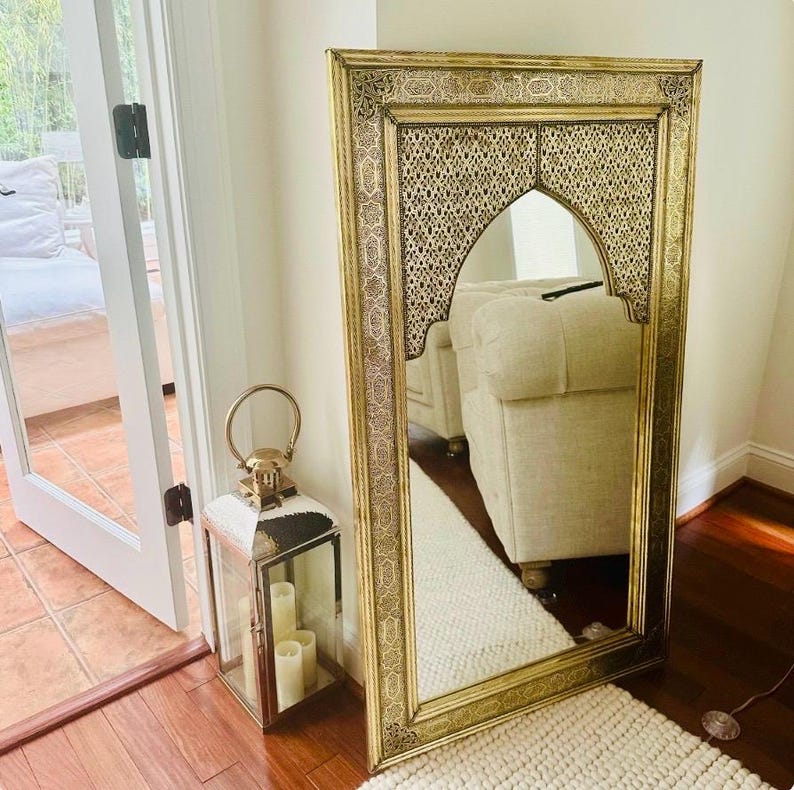 May include: A large, ornate gold-framed mirror with intricate detailing. The mirror has an arched top and is positioned against a white wall. A decorative lantern with candles sits nearby, adding to the room's ambiance. The mirror reflects a cream-colored sofa.