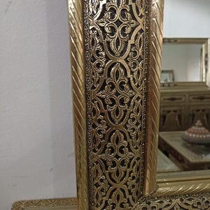 May include: A rectangular mirror featuring a wide, ornate gold frame with a dark, recessed background. The frame displays intricate, repeating floral and geometric patterns in relief, bordered by a twisted rope-like design on the inner and outer edges.
