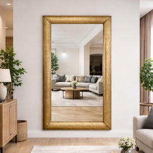 Peut inclure: Un grand miroir rectangulaire avec un cadre doré orné est accroché à un mur blanc. Le miroir reflète un salon avec un canapé, une table basse et des plantes. Le cadre présente un motif en relief détaillé.