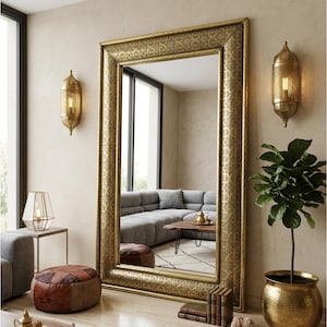 May include: A large, rectangular gold-framed mirror with an intricate pattern. The mirror reflects a living room with a grey sofa, coffee table, and ottoman. Two gold sconces are mounted on the wall. The room has a warm, inviting feel.