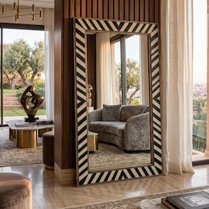 Peut inclure: Un grand miroir rectangulaire avec un cadre à chevrons noir et blanc. Le miroir reflète un salon avec un canapé gris, une table basse et une vue sur l'extérieur.