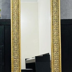 May include: A tall rectangular floor mirror featuring a wide gold-colored frame with an intricate, repeating openwork pattern and a twisted rope-style border. The reflective glass surface shows a clear view of a room with a black piano and bench.