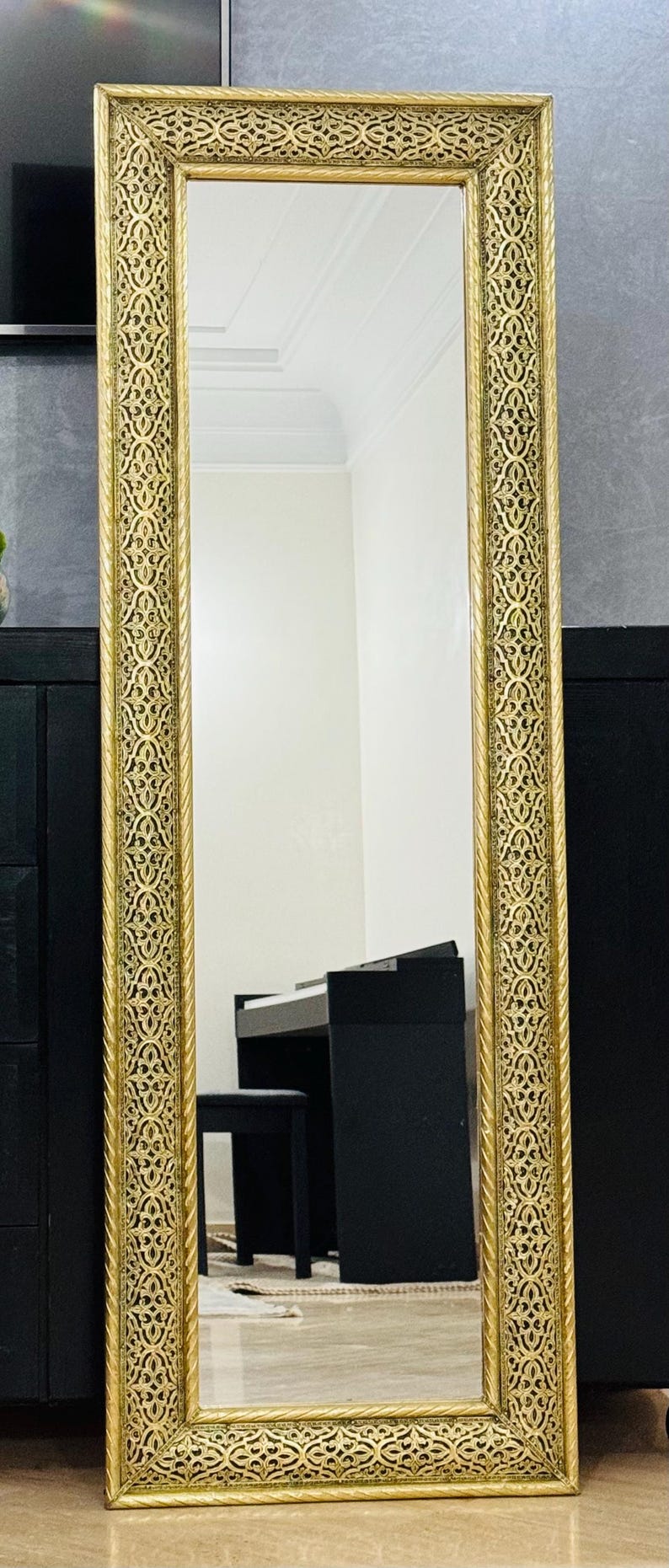 May include: A tall rectangular floor mirror featuring a wide gold-colored frame with an intricate, repeating openwork pattern and a twisted rope-style border. The reflective glass surface shows a clear view of a room with a black piano and bench.