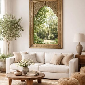 Salon avec un grand miroir doré orné au-dessus d'un canapé blanc. Des coussins beiges et une table basse ronde en bois avec des livres et des fleurs. Deux poufs en osier sont posés sur un tapis en jute.