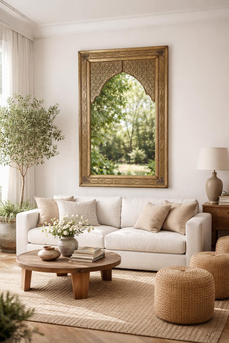 Salon avec un grand miroir doré orné au-dessus d'un canapé blanc. Des coussins beiges et une table basse ronde en bois avec des livres et des fleurs. Deux poufs en osier sont posés sur un tapis en jute.