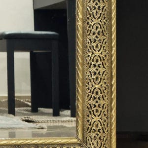 May include: A rectangular mirror featuring a wide, ornate gold-colored frame with a raised, intricate filigree pattern and a twisted rope-style border along the inner and outer edges, reflecting a black piano bench in the background.