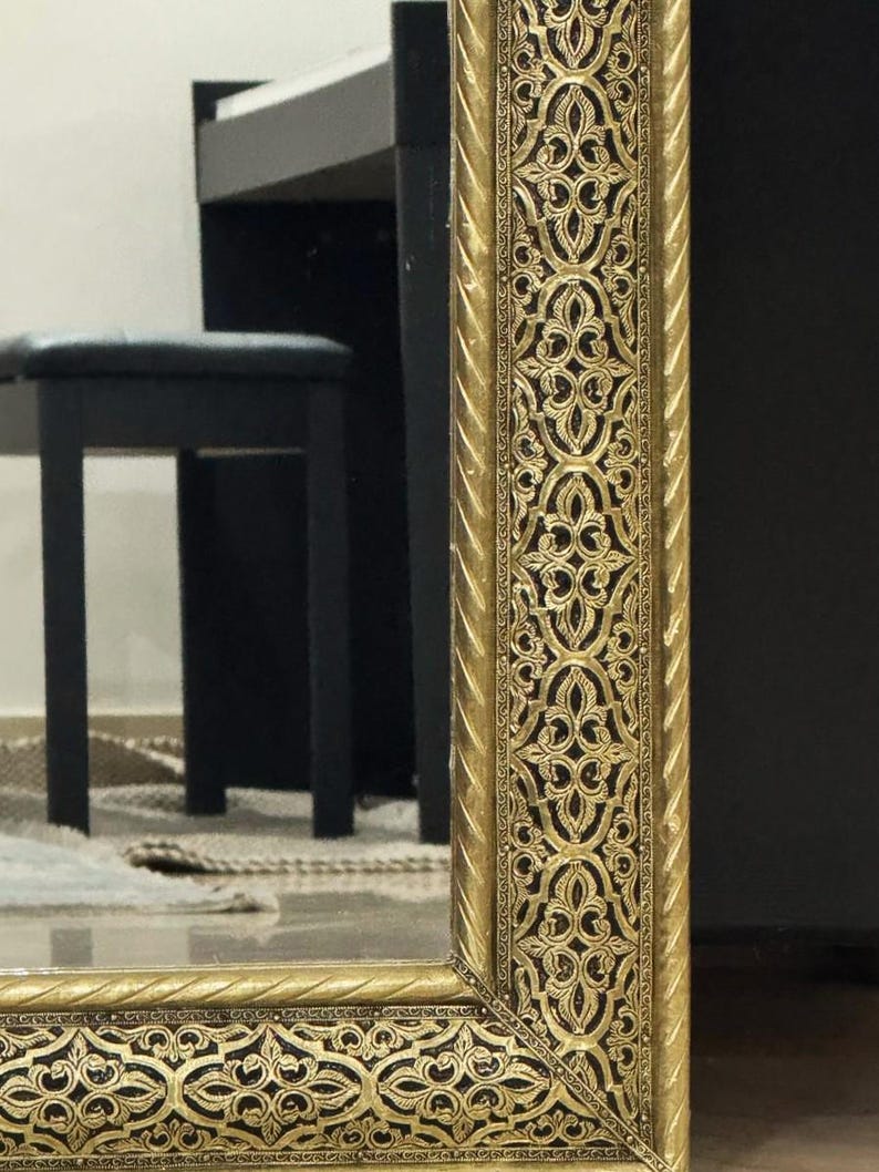 May include: A rectangular mirror featuring a wide, ornate gold-colored frame with a raised, intricate filigree pattern and a twisted rope-style border along the inner and outer edges, reflecting a black piano bench in the background.