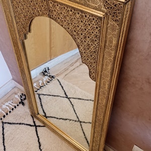 May include: A rectangular gold-colored mirror with an arched top. The frame features intricate, carved patterns. The mirror is angled against a pink wall and reflects a cream-colored rug with a black diamond pattern.