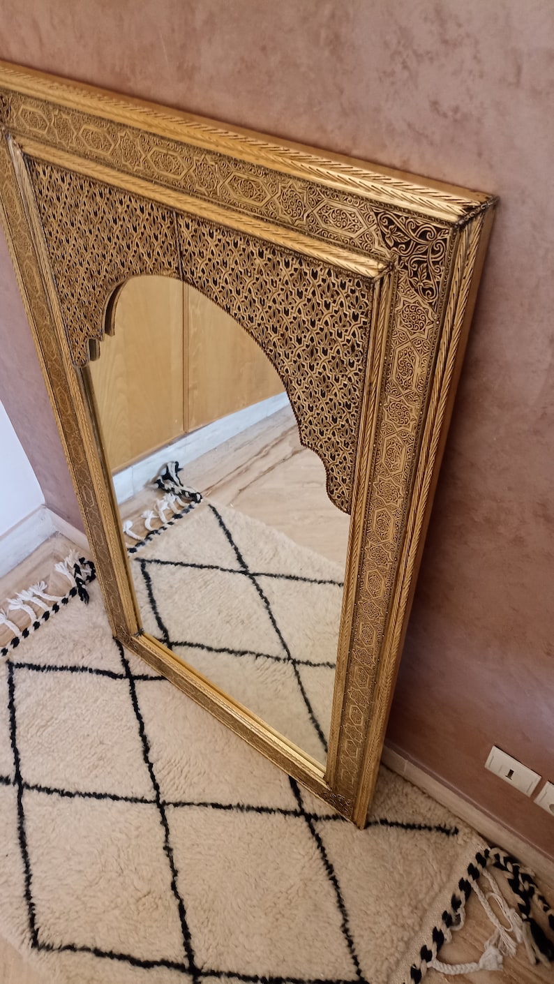 May include: A rectangular gold-colored mirror with an arched top. The frame features intricate, carved patterns. The mirror is angled against a pink wall and reflects a cream-colored rug with a black diamond pattern.