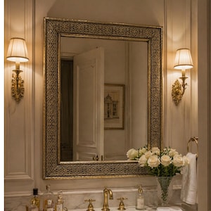 Peut inclure: Un grand miroir orné avec un cadre doré filigrané est suspendu au-dessus d'une coiffeuse en marbre. Deux appliques murales avec des abat-jours crème encadrent le miroir. Un vase de roses blanches est posé sur la coiffeuse.
