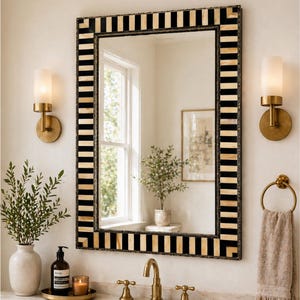 May include: Rectangular mirror with a black and cream striped frame, hanging above a bathroom sink. The mirror reflects a window and a framed picture. Two gold sconces flank the mirror, and a gold towel ring holds a beige towel. A vase of greenery and a soap dispenser are on the counter.