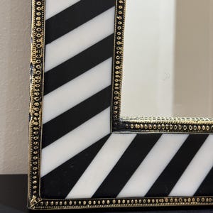 Pode incluir: Espelho decorativo com moldura listrada em preto e branco. A moldura tem uma borda dourada com pequenos detalhes circulares. O espelho é retangular e parece estar sobre uma superfície escura.