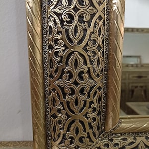May include: A rectangular mirror featuring a metallic gold frame with an intricate embossed floral pattern set against a dark, recessed background. The frame edges are finished with a twisted rope design, providing a textured border.