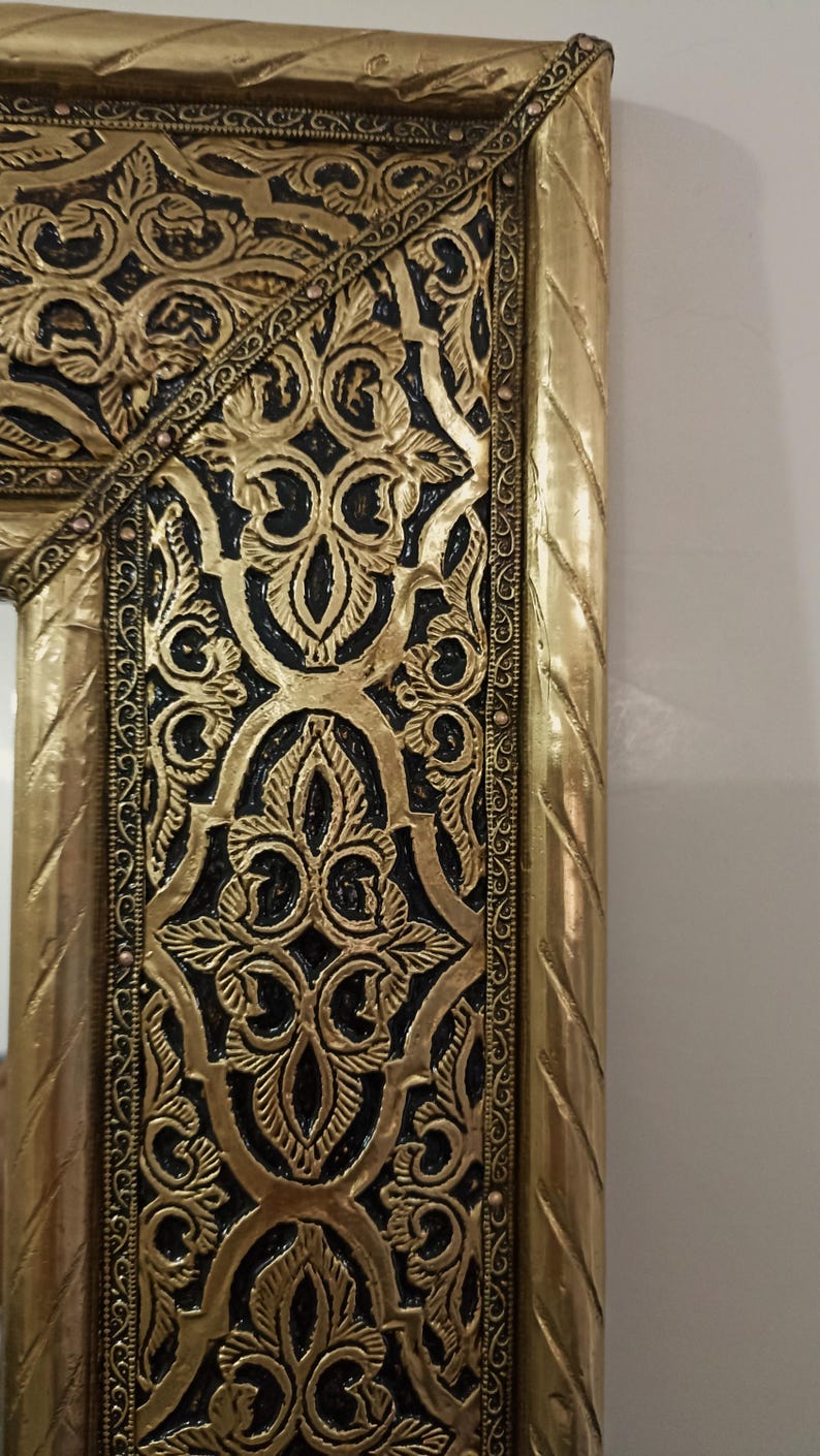 May include: A rectangular mirror frame featuring a metallic gold finish with intricate, raised floral and scrollwork patterns set against a recessed black background. The outer edge has a twisted rope texture, and the inner border is lined with small, evenly spaced metallic studs.