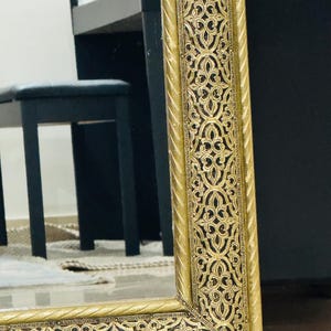 May include: A rectangular mirror featuring a wide, ornate gold frame with a raised, intricate filigree pattern set against a dark, recessed background. The frame edges are finished with a twisted rope-like texture, providing a decorative border.