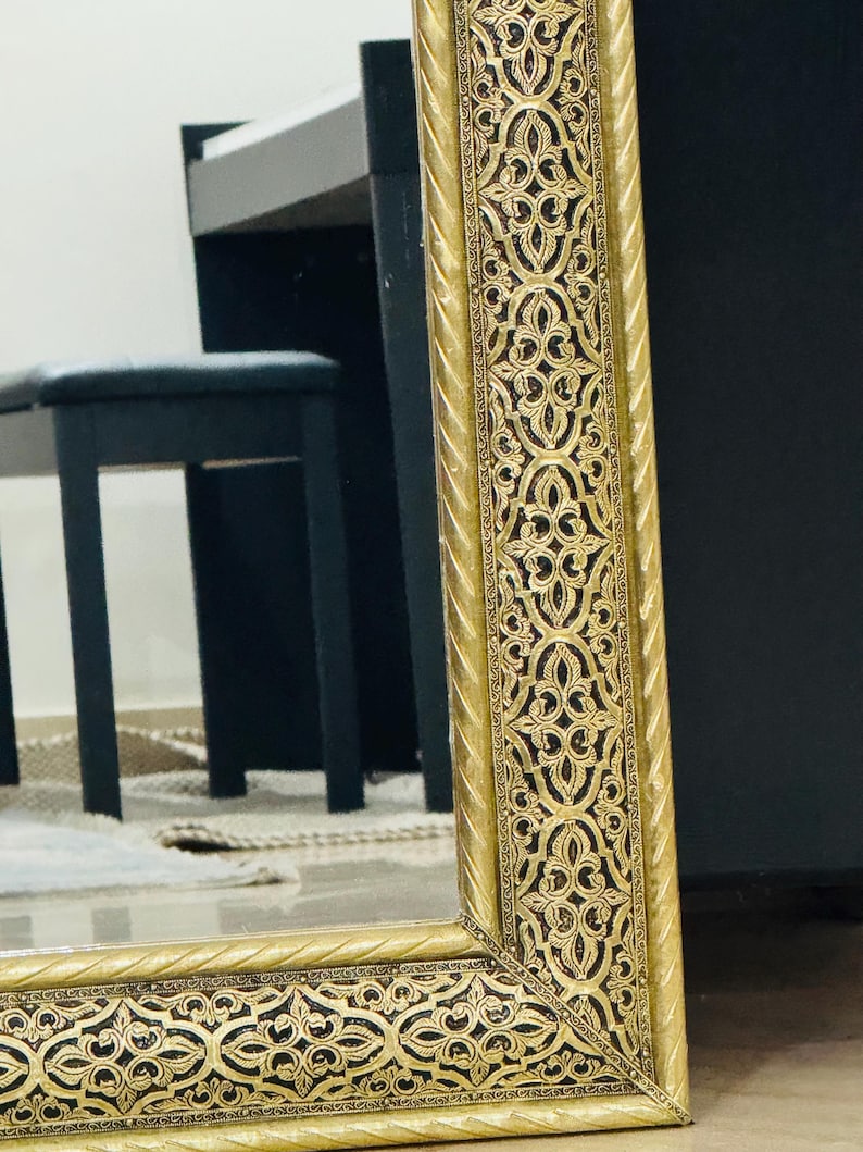 May include: A rectangular mirror featuring a wide, ornate gold frame with a raised, intricate filigree pattern set against a dark, recessed background. The frame edges are finished with a twisted rope-like texture, providing a decorative border.