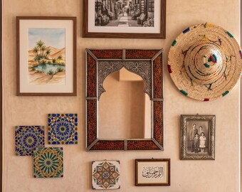 May include: A gallery wall arrangement featuring a central rectangular mirror with an ornate metal frame, a straw hat, three square patterned tiles, a framed watercolor of an oasis, a black and white street photograph, a family portrait, and calligraphy.