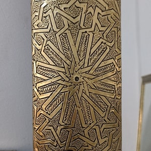 May include: A close-up of a gold-colored, rectangular object with an intricate geometric pattern. The design features repeating star and line motifs, creating a textured surface. The object has a dark, recessed background.