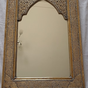 May include: Ornate gold-colored mirror with an arched top. The frame features intricate, repeating geometric and floral patterns. The mirror's surface reflects the surrounding environment, adding depth and dimension to the design.
