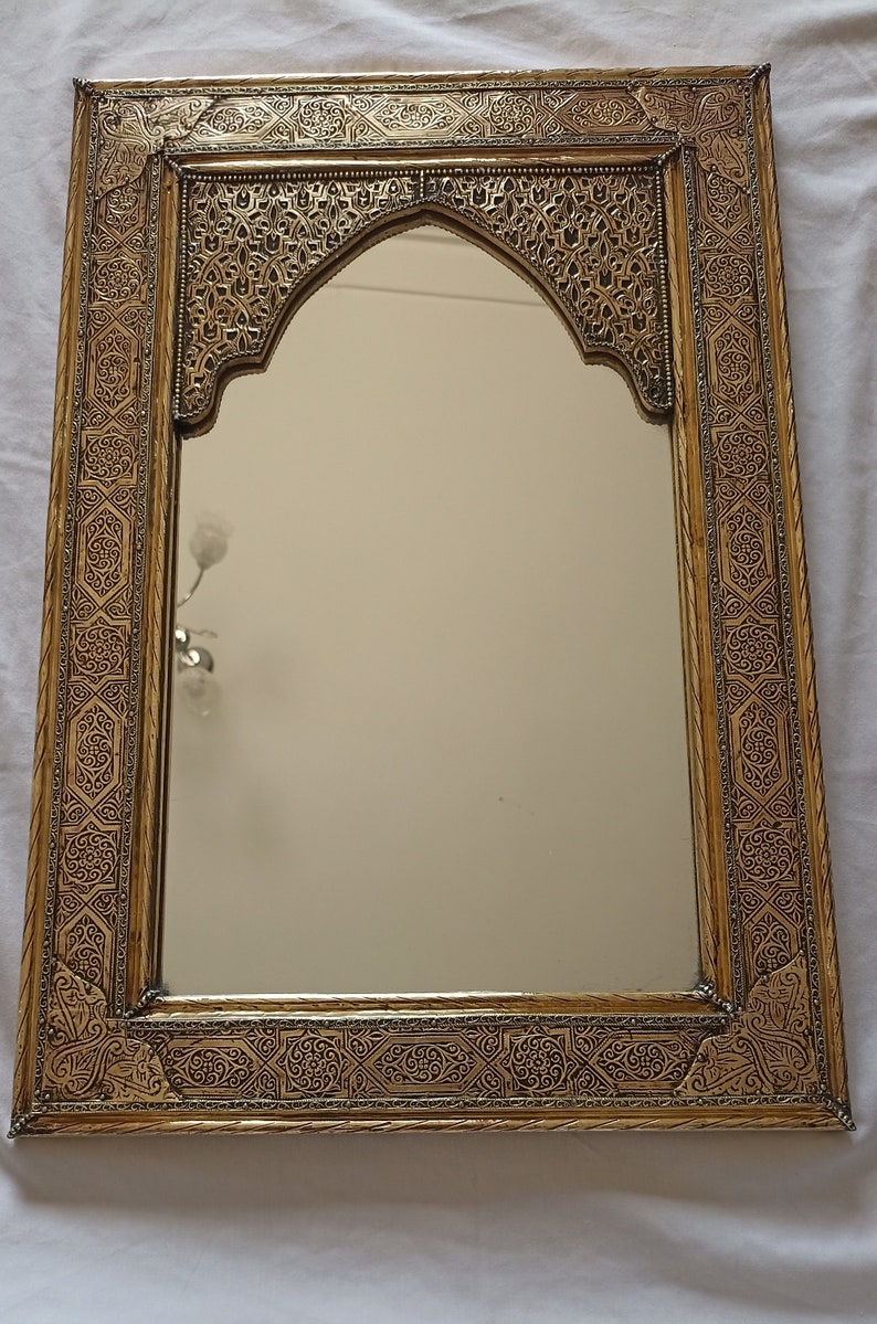 May include: Ornate gold-colored mirror with an arched top. The frame features intricate, repeating geometric and floral patterns. The mirror's surface reflects the surrounding environment, adding depth and dimension to the design.