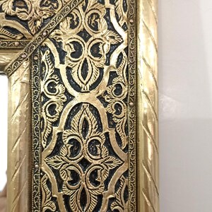 May include: A rectangular mirror frame featuring a metallic gold finish with intricate embossed floral and geometric patterns. The design includes a dark, recessed background that highlights the raised gold relief work and a twisted rope border.