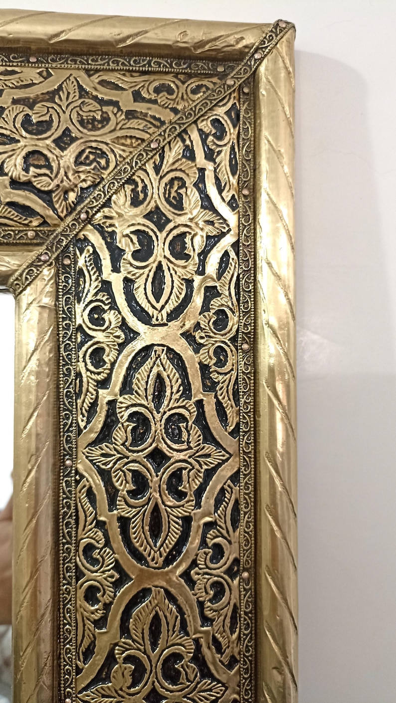 May include: A rectangular mirror frame featuring a metallic gold finish with intricate embossed floral and geometric patterns. The design includes a dark, recessed background that highlights the raised gold relief work and a twisted rope border.