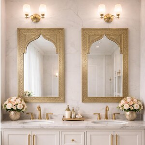 Pode incluir: Uma luxuosa bancada de banheiro com dois espelhos dourados, cada um com uma parte superior arqueada. A bancada possui torneiras douradas, pias brancas e uma bancada de mármore branco. Arranjos florais e arandelas completam o design elegante.