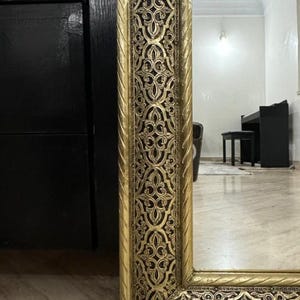 May include: A tall rectangular floor mirror featuring a wide, ornate gold frame with intricate, repeating floral-inspired carvings and a dark, recessed background. The frame edges are finished with a twisted rope-like texture in metallic gold.